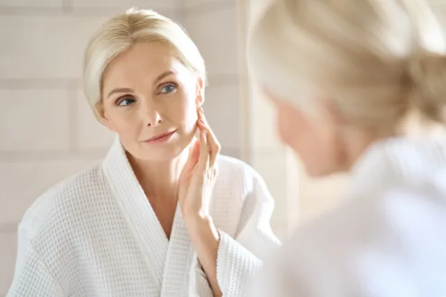 Headshot of mid age adult 50 years old blonde woman standing in bathroom after shower touching face, looking at reflection in mirror doing morning beauty routine.