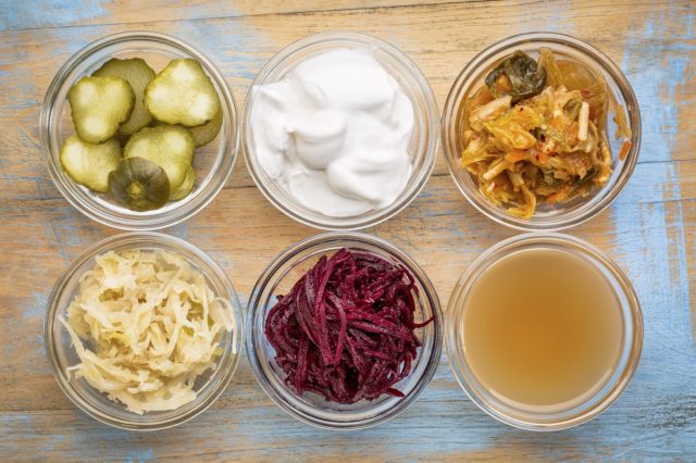 a set of fermented food great for gut health - top view of glass bowls against grunge wood: cucumber pickles, coconut milk yogurt, kimchi, sauerkraut, red beets, apple cider vinegar