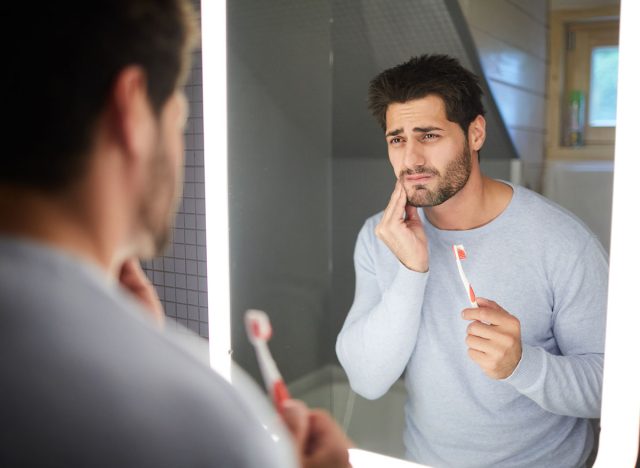 Sad frowning handsome brunette young man with beard touching cheek and looking into mirror in bathroom while getting bad tooth during cleaning teeth