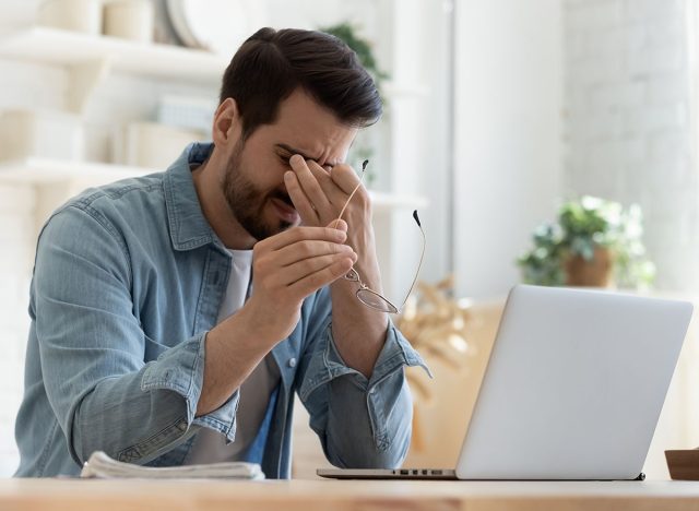 Tired young man feel pain eyestrain holding glasses rubbing dry irritated eyes fatigued from computer work, stressed man suffer from headache bad vision sight problem sit at home table using laptop