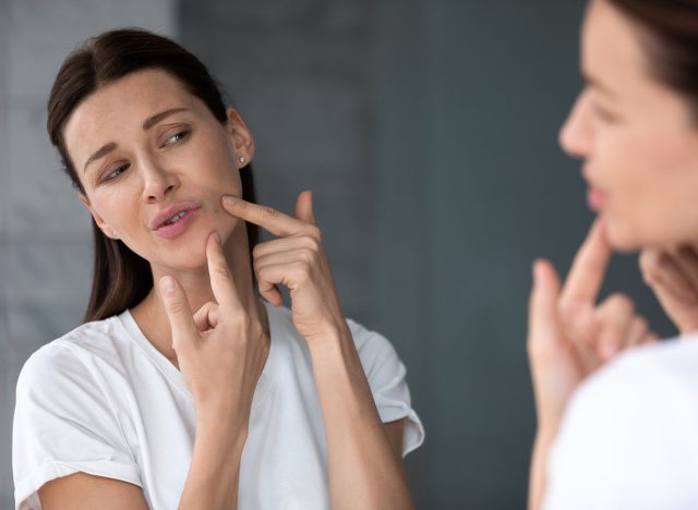 Over shoulder view upset female face reflected in mirror, woman touch face squeezes pimple caused by hormonal imbalance changes, stress or diet. Skin problem, jawline acne or new mole appears concept