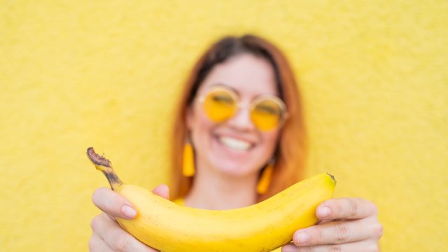 Beautiful red-haired woman in sunglasses holds a banana and fools around. Attractive European girl in a dress posing on a yellow background charmingly smiles.