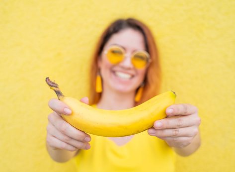 Beautiful red-haired woman in sunglasses holds a banana and fools around. Attractive European girl in a dress posing on a yellow background charmingly smiles.