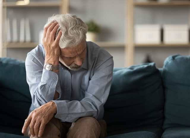 Frustrated unhappy middle aged mature man sitting on sofa, feeling depressed alone at home. Confused senior retired grandfather worrying about difficult life decision, copy space, old people solitude.