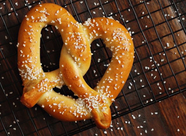 Freshly baked soft pretzel with generous sprinkling of coarse salt on wire cooling rack over rustic dark wood. Closeup from above.