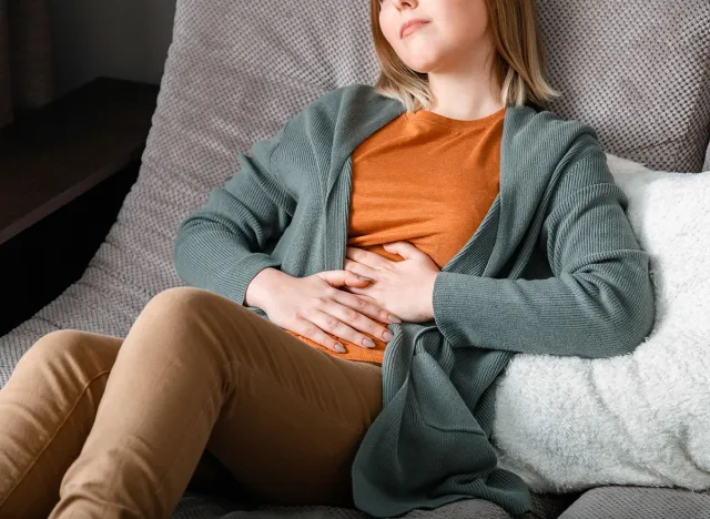 Young woman suffers, writhes in abdominal pain lying on couch in living room at home interior. Acute pain in bloating pms. Teenage girl with pain problems bowel disease