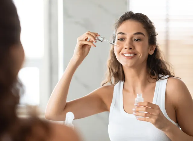 Facial Skincare. Attractive Female Applying Serum On Face Moisturizing And Caring For Skin Standing Near Mirror In Modern Bathroom Indoors. Beauty Routine. Skin Care Concept. Selective Focus