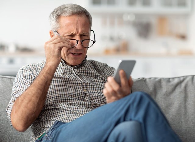 Poor Eyesight. Senior Man Squinting Eyes Reading Message On Phone Wearing Eyeglasses Having Problems With Vision Sitting On Couch At Home. Ophtalmic Issue, Bad Sight In Older Age Concept