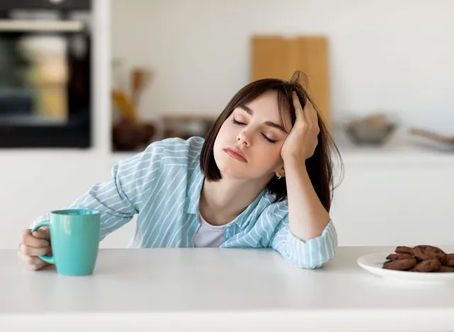 Sleepy young woman drinking coffee, feeling tired, suffering from insomnia and sleeping disorder. Sad female sitting in modern kitchen interior, empty space