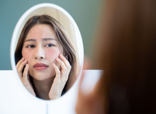 Asian young woman looking face skin in the mirror have a red rash on her face from cosmetic allergy