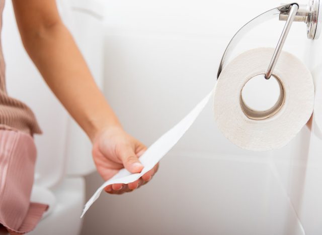 Closeup hand pulling toilet paper roll in holder for wipe, woman sitting on toilet she taking and tearing white tissue on wall to towel clean in bathroom, Healthcare concept