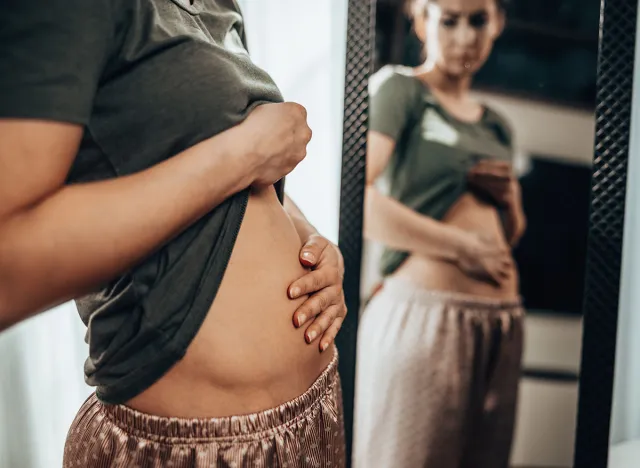 Unhappy young woman standing in front of a mirror and holding hands on her bloating stomach.