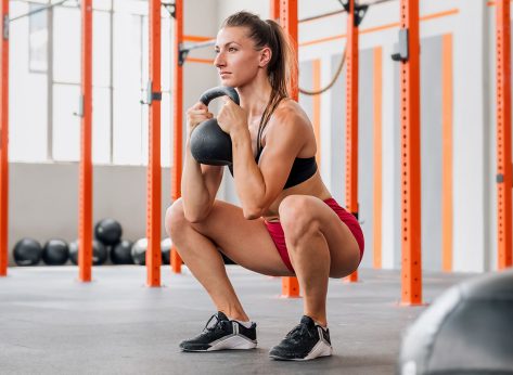 Determined female athlete looking away and doing goblet squat with heavy kettlebell during intense training in spacious light gym