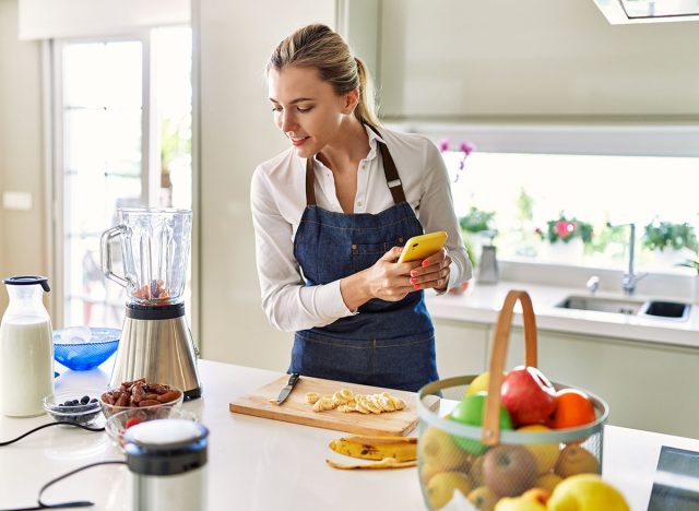 Young blonde woman using smartphone cutting banana at kitchen