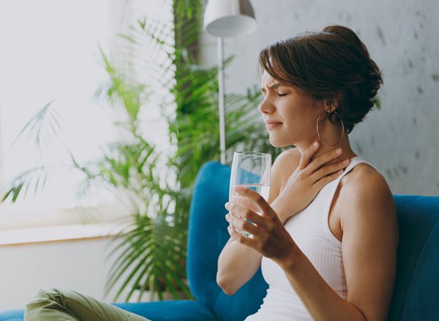 Side view young sad choked woman wears white tank shirt hold drink clear fresh pure still water from transparent glass hold neck sit on blue sofa stay at home flat spend time in living room indoors