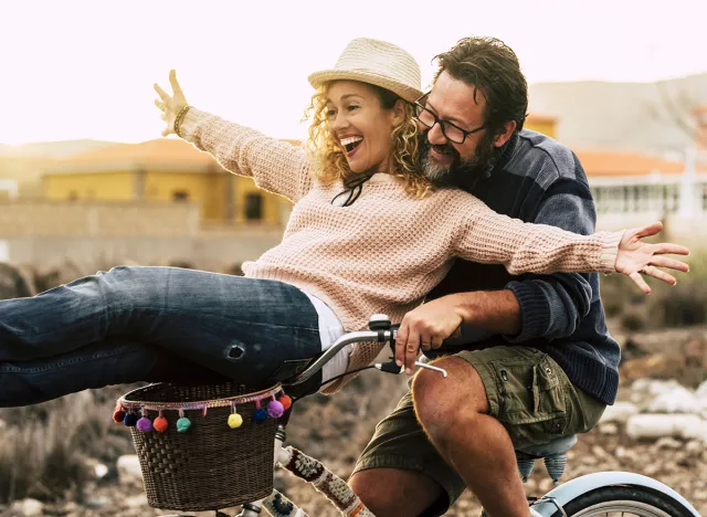 Happy couple enjoy outdoor leisure activity together carrying and using a bike and laughing a lot. Love and friendship with mature man and woman in youthful lifestyle. Concept of joyful and excitement