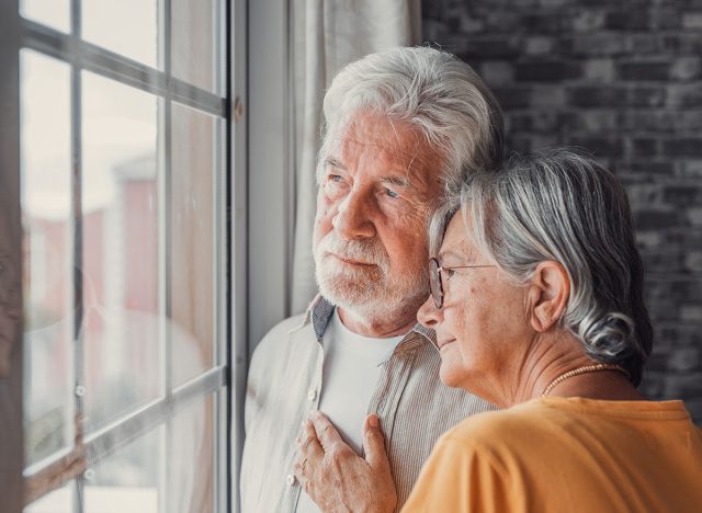 Pensive elderly mature senior man in eyeglasses looking in distance out of window, thinking of personal problems. Old woman wife consoling and hugging sad husband, copy space