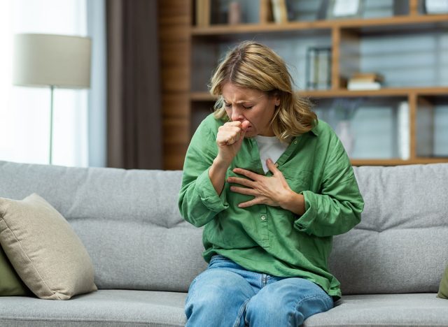 Senior beautiful woman sitting sick on sofa at home. He coughs, covers his mouth with his hand, holds his chest. Feels pain, suffers from asthma, allergies, flu, cold.