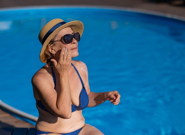 Portrait of an old woman in a straw hat, sunglasses and a swimsuit applying sunscreen to her face while relaxing by the pool.