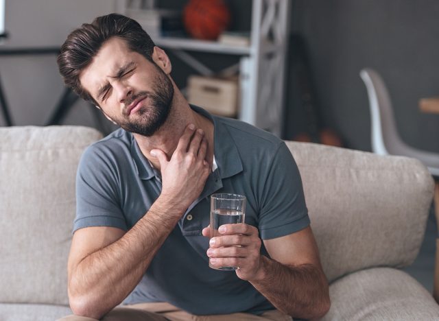 Terrible pain in his throat. Frustrated handsome young man touching his neck and holding a glass of water while sitting on the couch at home