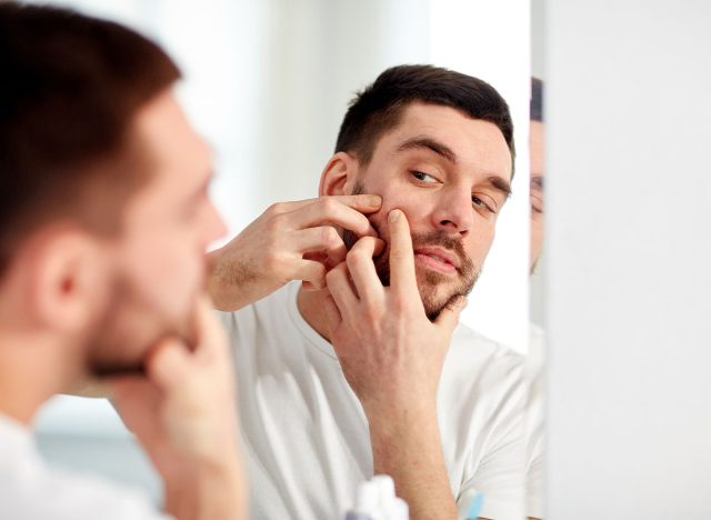 beauty, hygiene, skin problem and people concept - young man looking to mirror and squeezing pimple at home bathroom