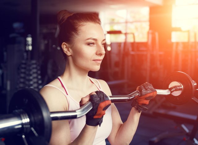 Young beautiful woman doing biceps curl with EZ curl bar in a gym. Athletic girl doing workout in a fitness center with sunset beams in the window