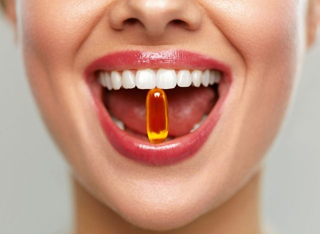 Vitamins And Food Supplements. Close Up Of Beautiful Woman Opened Mouth Holding Fish Oil Pill In White Teeth. Smiling Girl Holding Capsule With Omega-3 Between Teeth. Healthy Diet Nutrition Concept