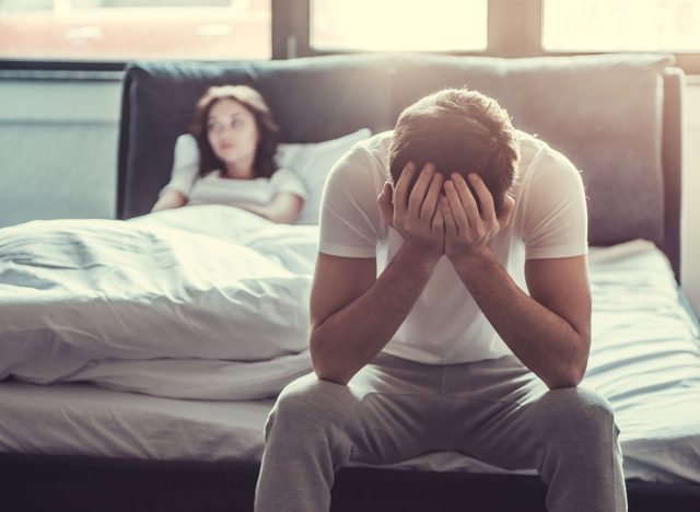 We have a problem. Upset young man sitting on the edge of the bed and holding his head against the background of a girlfriend, lying in bed.