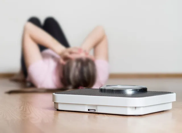 Weight loss fail concept. Scale and depressed, frustrated and sad woman lying on floor holding head and covering face with hands.