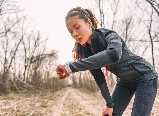 Sport watch run woman checking smartwatch tracker. Trail running runner girl looking at heart rate monitor smart watch in forest wearing jacket sportswear. Female athlete jogger training in woods.