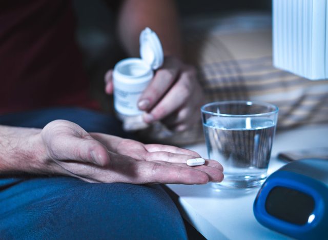 Man sitting on bed about to take sleeping pill or night medicine. Suffering from insomnia. Holding tablet and bottle in hand. Glass of water on nightstand in bedroom.