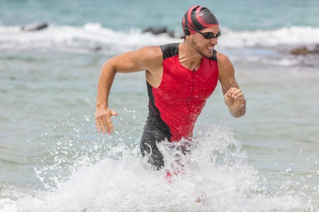 Triathlon swimming man. Male triathlete swimmer running out of ocean finishing swim race. Fit man ending swimming sprinting determined out of water in professional triathlon suit training for ironman.