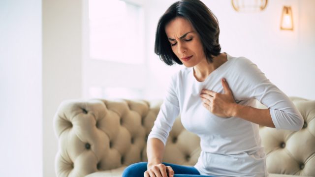 Close-up photo of a brunette woman who is sitting on a couch with her eyes closed and her left hand touching her heart area.