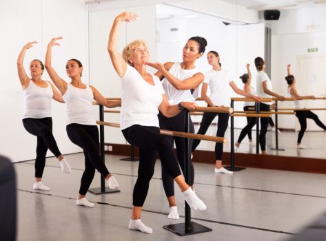 Group of women engaged in ballet in a dance studio perfoms an exercise near the ballet barre, where the choreographer helps ..to coordinate the movements correctly