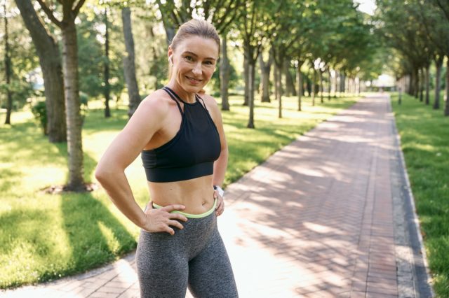 Sporty mature woman in sportswear and earphones looking at camera while jogging in a green park on a sunny summer day