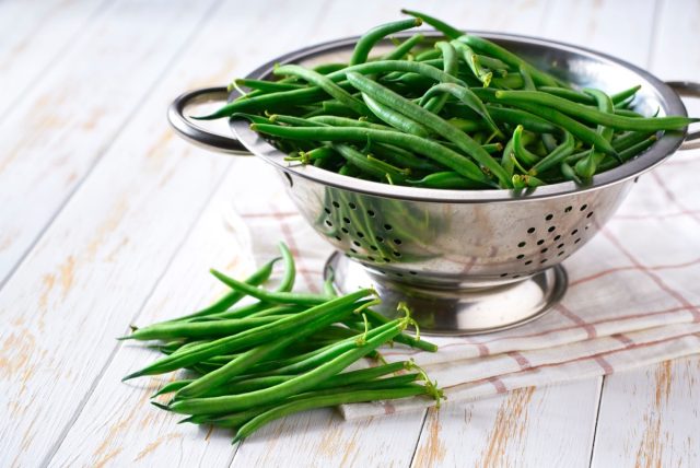 Green beans in a steel strainer sieve metal on a white wooden table, selective focus.