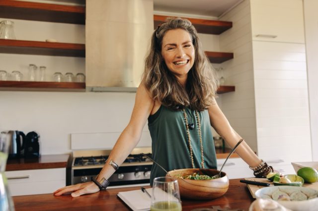 Happy vegan woman smiling at the camera while preparing a plant-based meal. Mature woman following an organic recipe in her kitchen. Healthy senior woman eating clean at home.