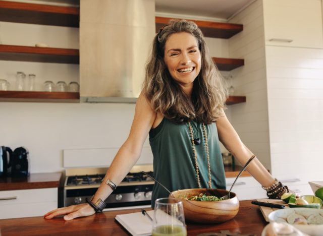 Happy vegan woman smiling at the camera while preparing a plant-based meal. Mature woman following an organic recipe in her kitchen. Healthy senior woman eating clean at home.