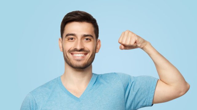 Young strong sporty athletic man in casual t-shirt, showing biceps after training in gym, isolated on blue background
