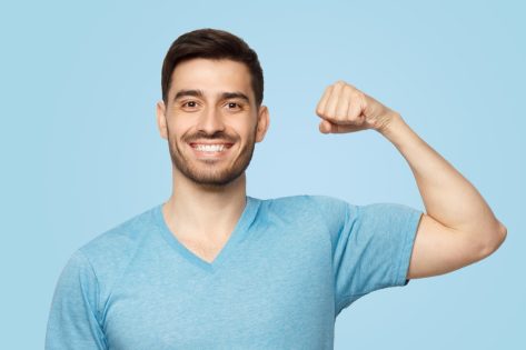 Young strong sporty athletic man in casual t-shirt, showing biceps after training in gym, isolated on blue background