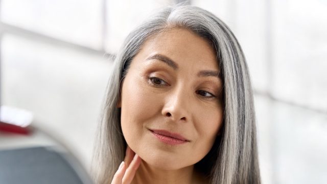 Portrait of gorgeous happy middle aged mature asian woman, senior older 50s lady pampering touching face looking at herself at mirror indoors