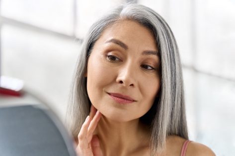 Portrait of gorgeous happy middle aged mature asian woman, senior older 50s lady pampering touching face looking at herself at mirror indoors