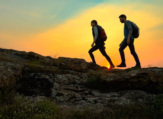 Couple of Young Happy Travelers Hiking with Backpacks on the Beautiful Rocky Trail at Warm Summer Sunset. Family Travel and Adventure Concept.