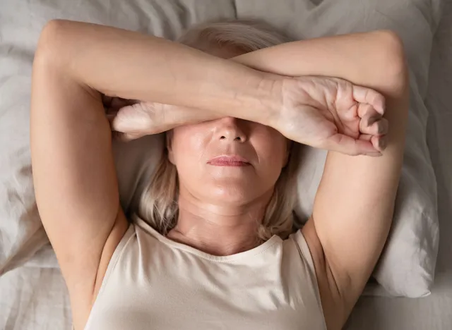 Sleepless mature woman suffering from insomnia close up, lying in bed, older female covering eyes with hands, trying to sleep, nightmares or depression, feeling headache or migraine