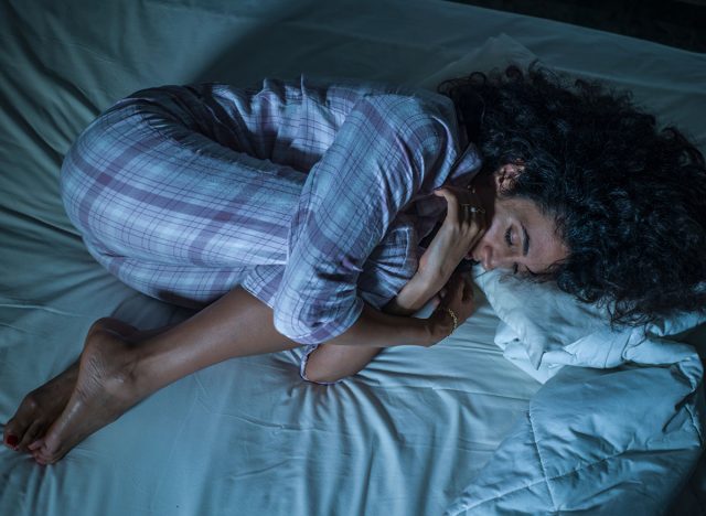 dramatic night lifestyle portrait of young sad and depressed latin woman with curly hair sleepless in bed suffering excruciating period pain holding her belly in fetal position