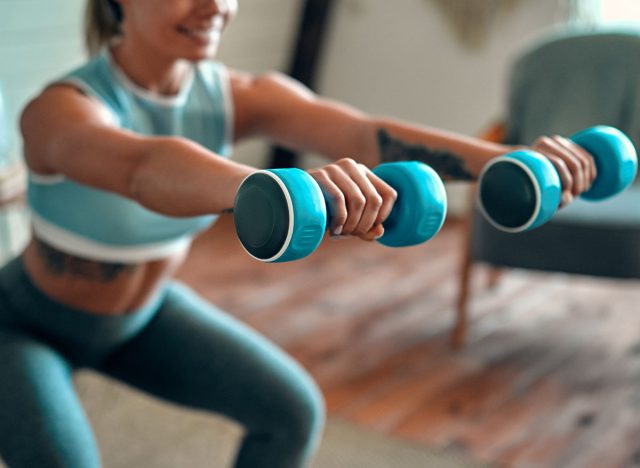 Blurred and cropped image of a beautiful young athletic girl in leggings and top crouches with dumbbells at home. Sport, healthy lifestyle.