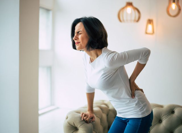 Chronic back pain. Adult woman is holding her lower back, while standing and suffering from unbearable pain.