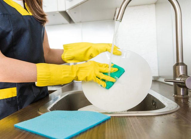 A young woman in yellow gloves washes dishes with a sponge in the sink. House professional cleaning service.