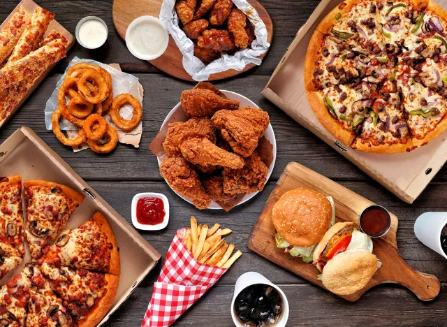 Buffet table scene of take out or delivery foods. Pizza, hamburgers, fried chicken and sides. Above view on a dark wood background.