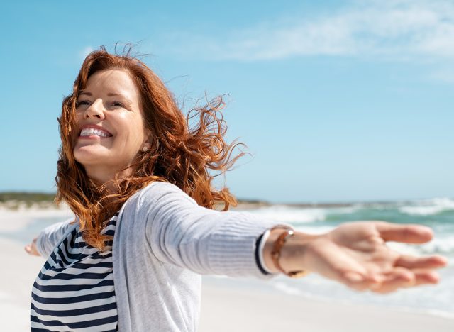 Happy mature woman with arms outstretched feeling the breeze at beach. Beautiful middle aged woman with arms up dancing on beach. Mid lady feeling good and enjoying freedom at sea, copy space.